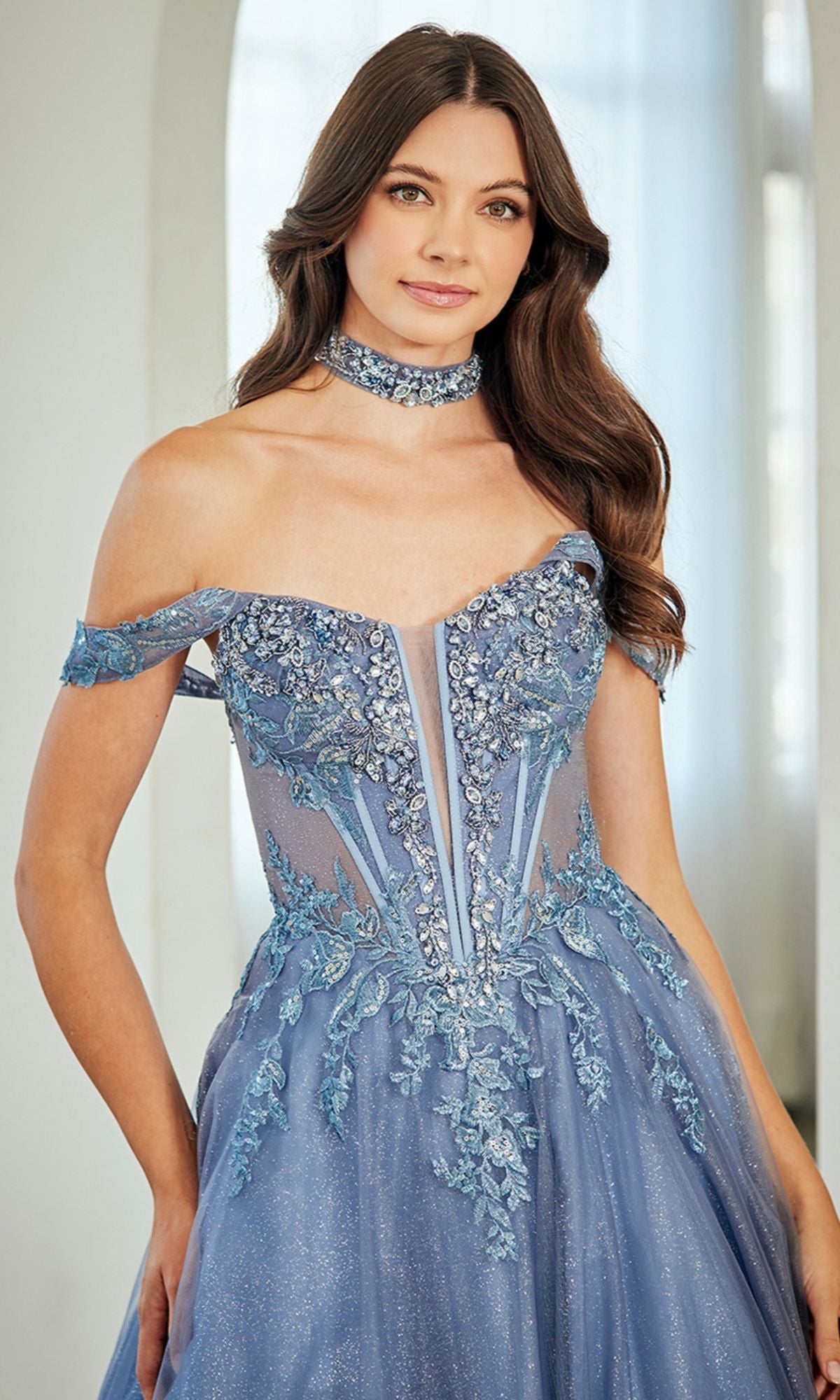 A woman poses indoors, smiling slightly in the Amelia Couture 8065 long blue off-the-shoulder prom dress featuring floral embroidery, a corset bodice, and a matching choker.